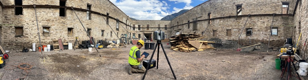 Wide view of a technician with a 3D laser scanner on a tripod in a historic stone courtyard under renovation, with safety gear and construction materials
