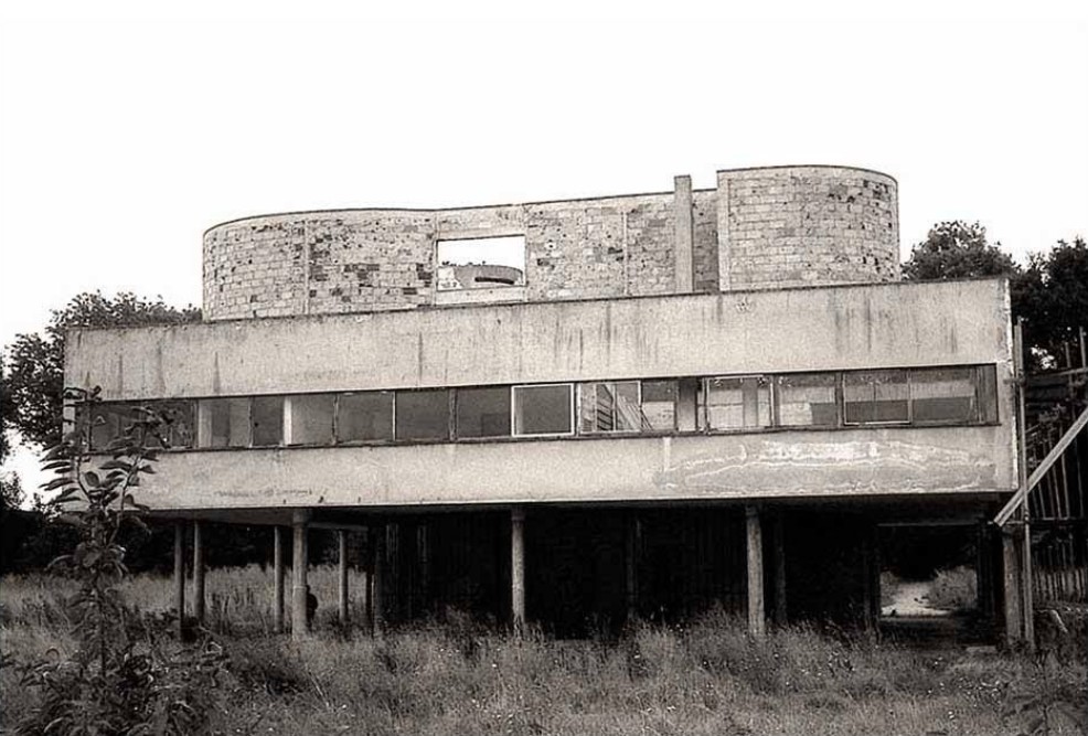 Black-and-white photograph of Le Corbusier’s Villa Savoye: weathered modernist facades on pilotis, ribbon windows, and overgrown foreground—illustrating architectural neglect and decay